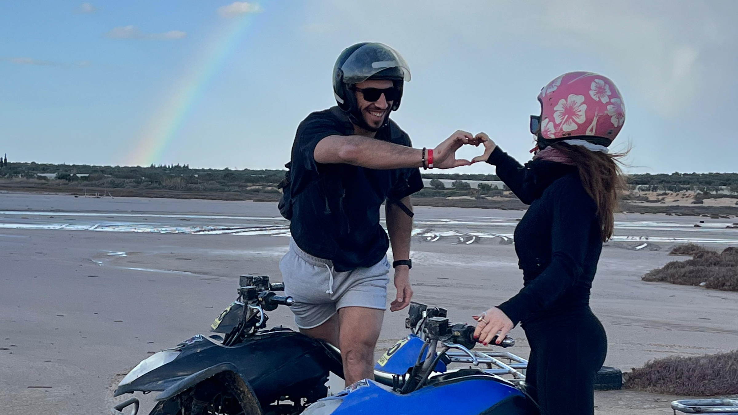 Juan et Emy forment un cœur avec leurs mains, en extérieur, sous un ciel de fin de journée