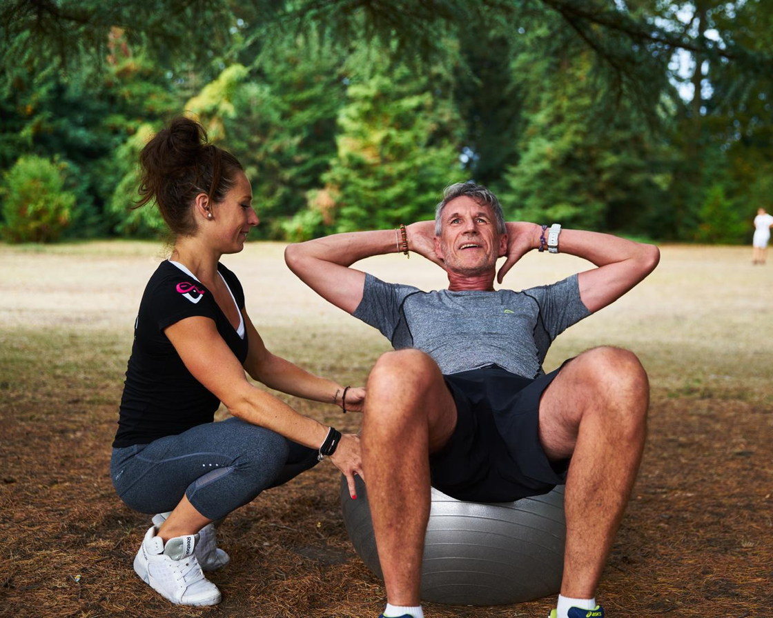 Emy assise dans l'herbe tient les jambes d'un client pendant une série d'abdominaux, tous les deux souriants