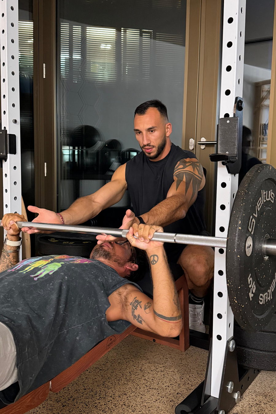Juan, debout derrière le rack, tient la barre à deux mains pendant qu'un client termine un développé couché