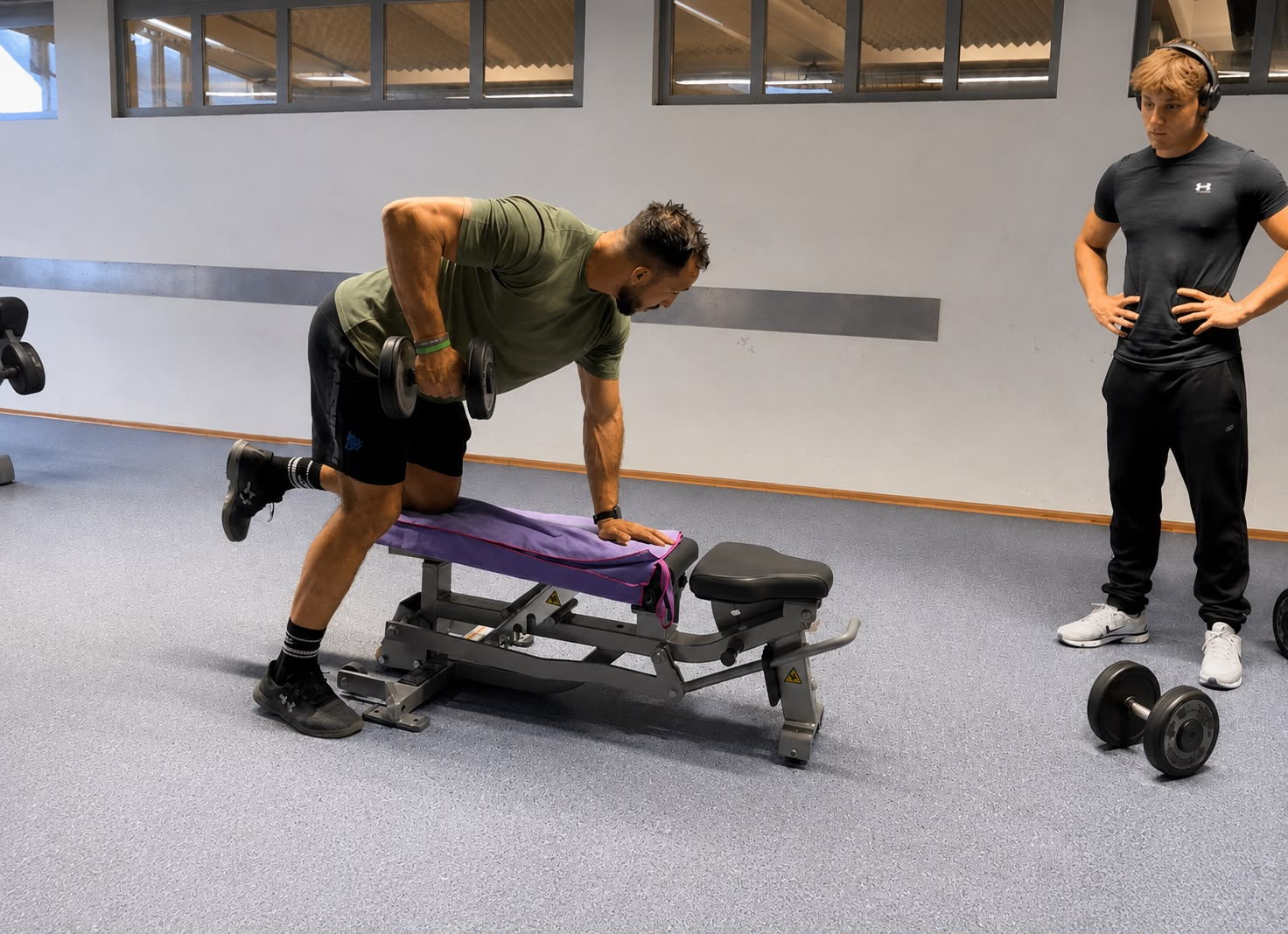 Juan exécute un rowing haltère à un bras, en appui sur un banc ; une autre personne suit le mouvement debout à droite